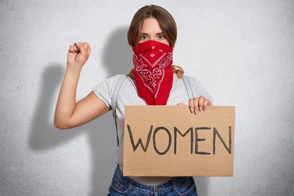 Feminism concept. Self confident young woman feminist protects womens rights, takes part in protest, keeps hands in fist raised, wears red bandana and grey t shirt, stands indoor, being serious