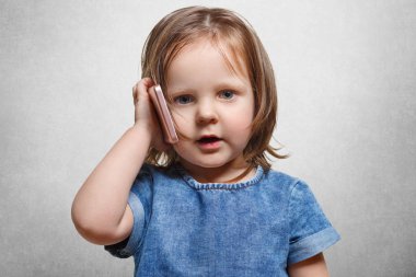 Sevimli küçük erkek çocuk, son moda saç modeli vardır, denim moda elbise giyer, modern akıllı fon beyaz beton artalan üzerinde izole iletişim için kullanır. Çocukluk ve teknolojileri kavramı