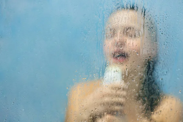 Relaxed carefree woman washes herself in douche, sings loudly, pretends to be successful talented singer, enjoys loneliness at home, poses behind blurred glass with water drops and copy space for text