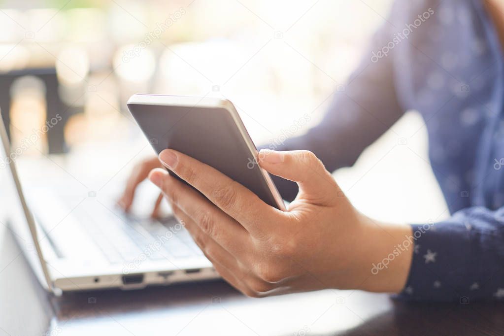 Modern technology and lifestyle concept. A close-up of woman`s hands holding smartphone and typing something at her laptop. A woman having dinner break at cafe using free internet connection. 