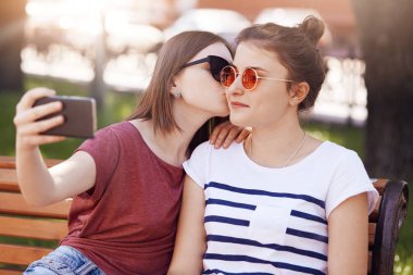Kadın Lezbiyenler parkta boş zaman zevk, akıllı telefon selfie yapmak için poz. Güzel erkek onun en iyi arkadaşı yanak içinde öpüyor, aşk ifade eder, doğru ilişkileri vardır. Leisute zaman kavramı