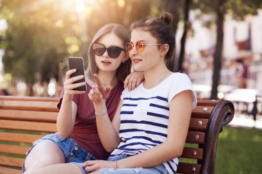 Trendy tonları, akıllı telefon, dikkatle bakar güzel kadın gençler online alışveriş yapmak, yeni yaz kıyafeti seçin, parkta bankta poz, güneşli sıcak havanın tadını çıkarmak. Rekreasyon kavramı