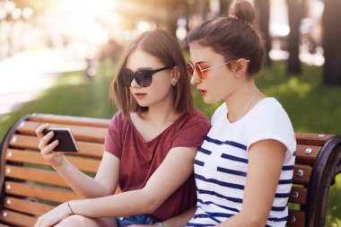Gündelik yaz giysileri, cep telefonu, içine bak attentivwly oldukça iki kız öğrencilerin yatay atış bankta intersting seri, recreat biri için beklemek süre dikkat et. İnsanlar, park, dinlenme