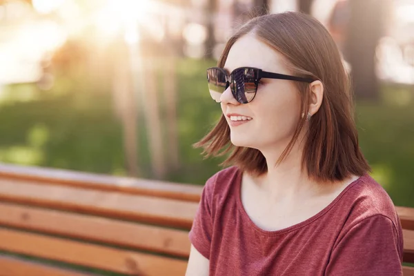 トレンディなヘアスタイルで肯定的な若い素敵な女性おしゃれな色合いを身に着けている 公園のベンチに座って 夏季に晴天を楽しんで 距離に見えます 陽気な学生の屋外撮影 — ストック写真
