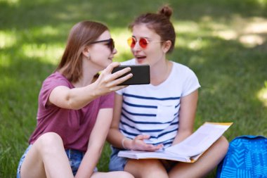 Gençlik ve teknoloji kavramı. Pozitif en iyi genç arkadaşlar yakından oturup, kitap okumak ve selfie portre yapmak için poz, modern akıllı telefon üzerinde odaklanmak, yeşil doğa arka planı poz
