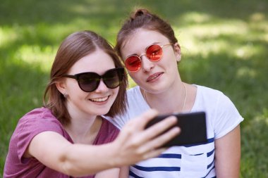 Yakın bobin saç kesimi ile gülümseyen kadın genç bir kadeh, trendy siyah tonları giyer, pozlar yakın arkadaşı olun selfie modern cep telefonuyla fotoğraf sosyal ağlar paylaşacak
