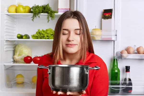 Pleased female holds saucepan, smells pleasant oudour, cooked delicious soup with vegetables, stands at kitchen near opened fridge. Female vegeterian satisfied with new recipe of dish. Cooking concept