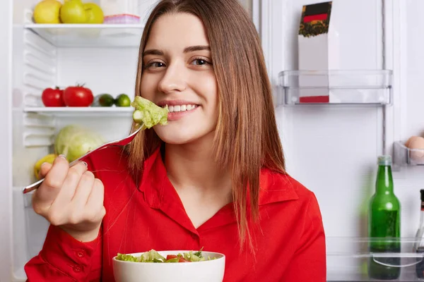 Lovely young woman eats fresh vegetable salad, tastes cabbage on fork, poses against opened refrigerator full of fruits and vegetables, has positive expression, keeps to diet. People and food concept