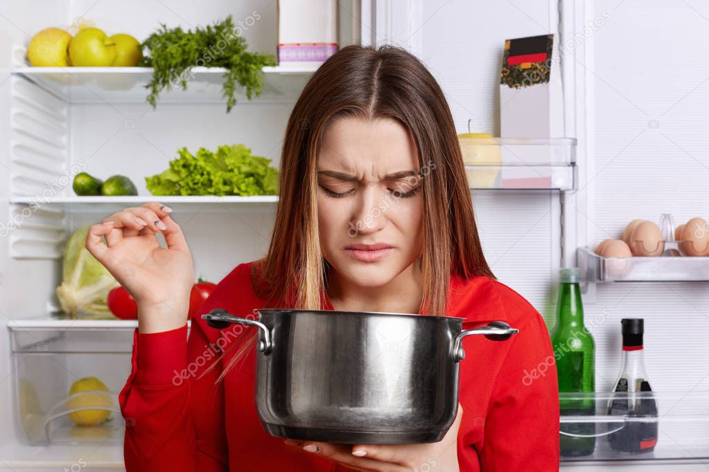 Pretty woman holds saucepan, feels unpleasant stink as there is spoiled dish, wears red blouse, stands near opened fridge, going to cook fresh dish. People, cooking, culinary and nutrition concept