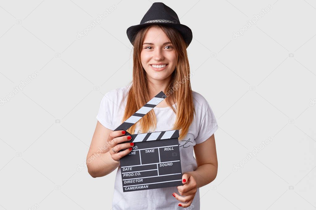 Film production concept. Pleased Caucasian female holds movie clapper, wears fashionable hat and casual white t shirt, isolated over white background. Beautiful actress participates in shooting movie