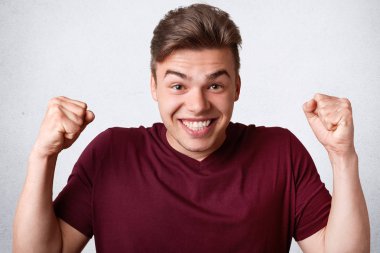 Kafkas erkek başarısını kutluyor gibi yumruklarını clenches çok memnun, iyi ruh hali, pozlar beyaz beton duvara olmak geniş gülümsemesi var. Mutlu pozitif genç adam jestleri, ifade mutluluk