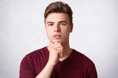 Yakın çekim yakışıklı hipster adam portresi güvenle kameraya benziyor, çene tutar, sağlıklı cilt ve modaya uygun saç modeli, hayatında bir şey hakkında contemplates, beyaz beton duvara pozlar