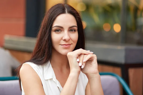 stock image Close up shot of pretty brunette young female with dark hair, healthy skin, holds hands under chin, looks mysteriously at camera, has pleasant talk with friend, poses against blurred background