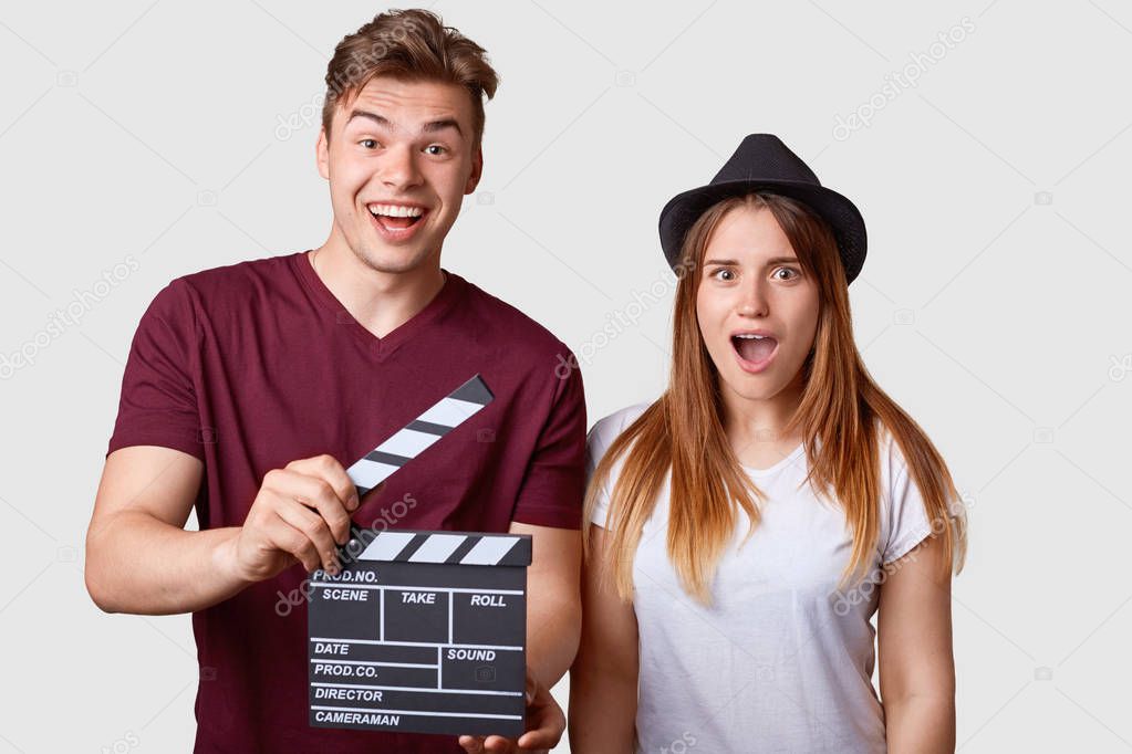 Horizontal portrait of shocked young successful female actress and her producer holds clapperboard, has cheerful expression, creat new production of film, stand close to each other, isolated on white