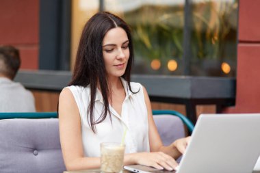 Koyu saçlı, dizüstü bilgisayar, klavye bilgi iyi görünümlü genç erkek fotoğrafı uzaktan çalışır, kokteyl içer, kahvehane iç karşı boş zaman harcıyor. Teknoloji kavramı