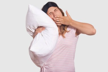 Yorgun öğrenci stüdyo çekim uzun hazırlık sınavı için sonra yatmak istiyor, esnemek, palm ile ağız kapsar, baş, rahat t gömlek tutar yastık, beyaz arka plan üzerinde izole üzerinde blindfellen giyer