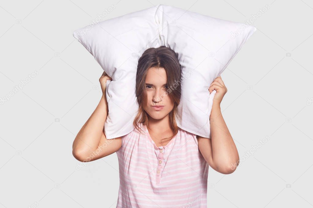 Irritated female cant sleep because of noise, tries to cover ears with pillow, has sleepless night as neighbours interrupt with noisy party, dressed in nightwear. People and insomnia concept