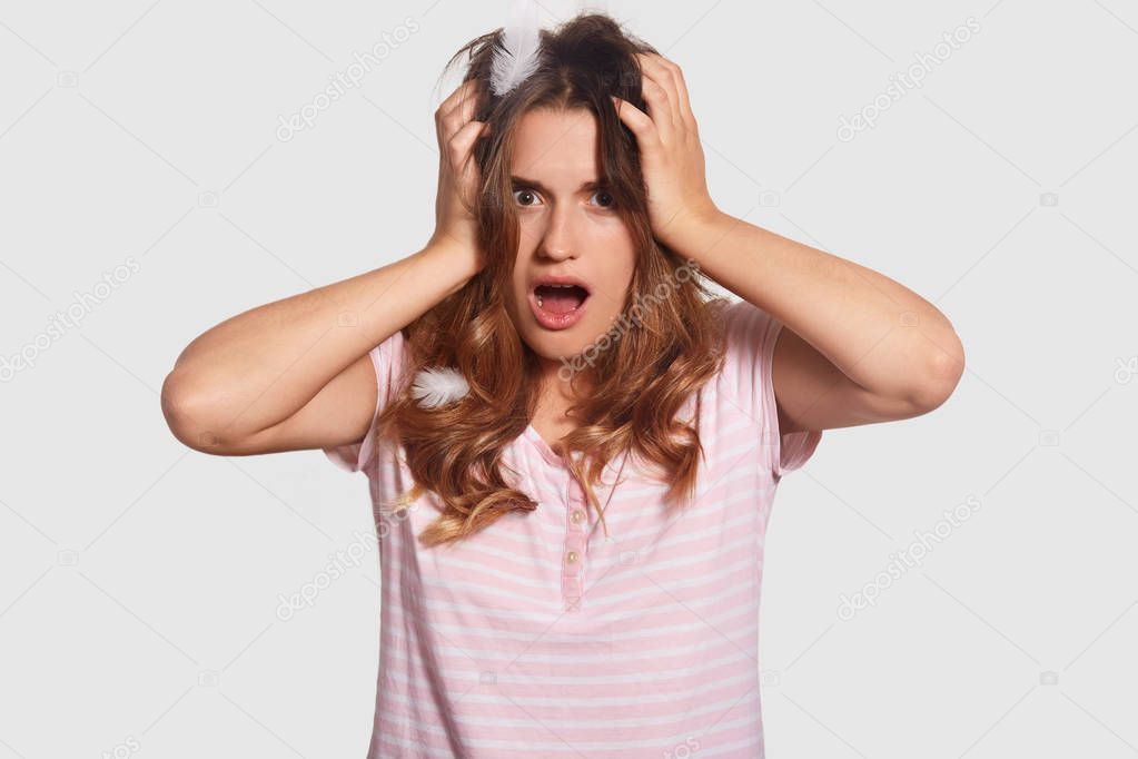 Shocked young pretty European woman has headache, keeps hands on head, overslept work, looks with terrified expression, wears nightclothes, isolated over white background. Omg, its too late!