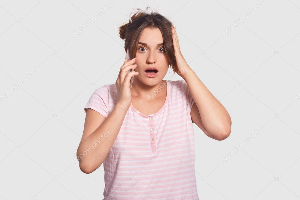 Surprised female has unexpected talk via cell phone, looks with terrific expression, dressed in nightclothes, opens mouth, speaks with friend, isolated over white background. People, reaction concept