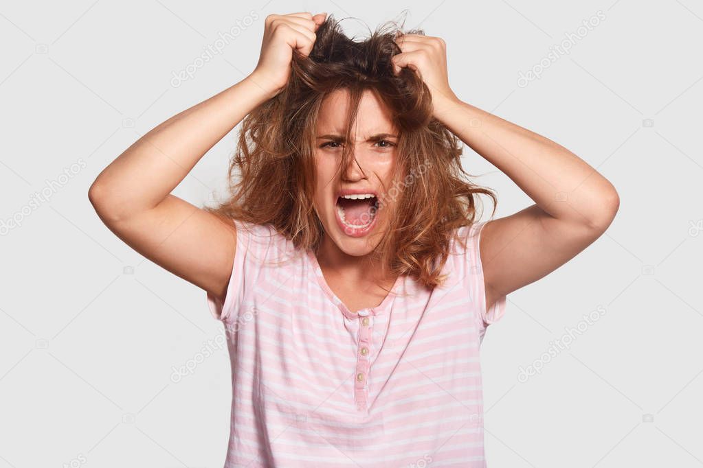 Depressed irritated young female has mess on hair, opens mouth widely, shouts angrily, doesnt want get up, fed up of daily routine, wants good rest and sleep, wears casual clothes, feels peevish