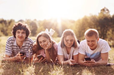 Teknoloji, gençlik ve boş zaman zaman kavramı. Mutlu dört arkadaş cep telefonu kullanımı, alanın, güneş ışığı ile arka plan bulanık yeşil çim üzerinde yalan. Sınıf cep arkadaşlarıyla piknik mesaj var