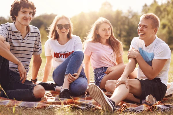 Outdoor shot of happy young friends have fun together, sit close to each other, tell anecdotes, enjoy warm sunny summer weather, have good rest, unforegattable day. Four teenagers on picnic.