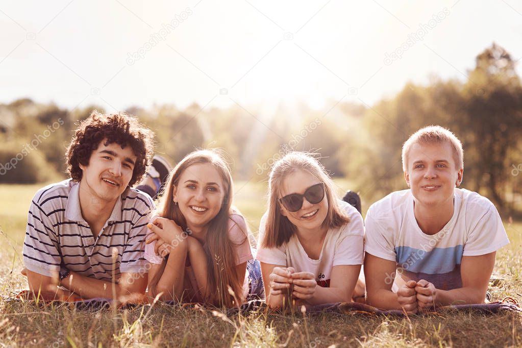 Smiling friends have happy expressions, lie on plaid on ground, rejoice sunny day, look at camera, spend free time together. Teenage groupmates celebrate starting summer holidays, have picnic
