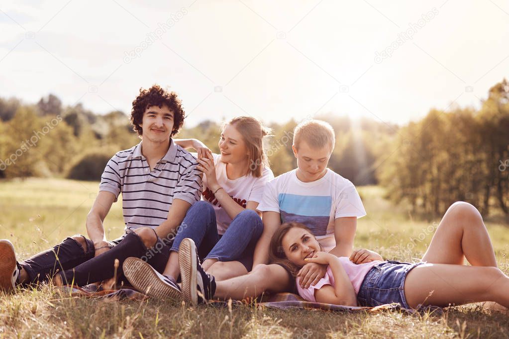 Outdoor shot of cheerful youth couples enjoy togetherness and spare time during summer day or weekend, sit closely to each other, admire beautiful nature, have pleasant talk. Teenagers and relations
