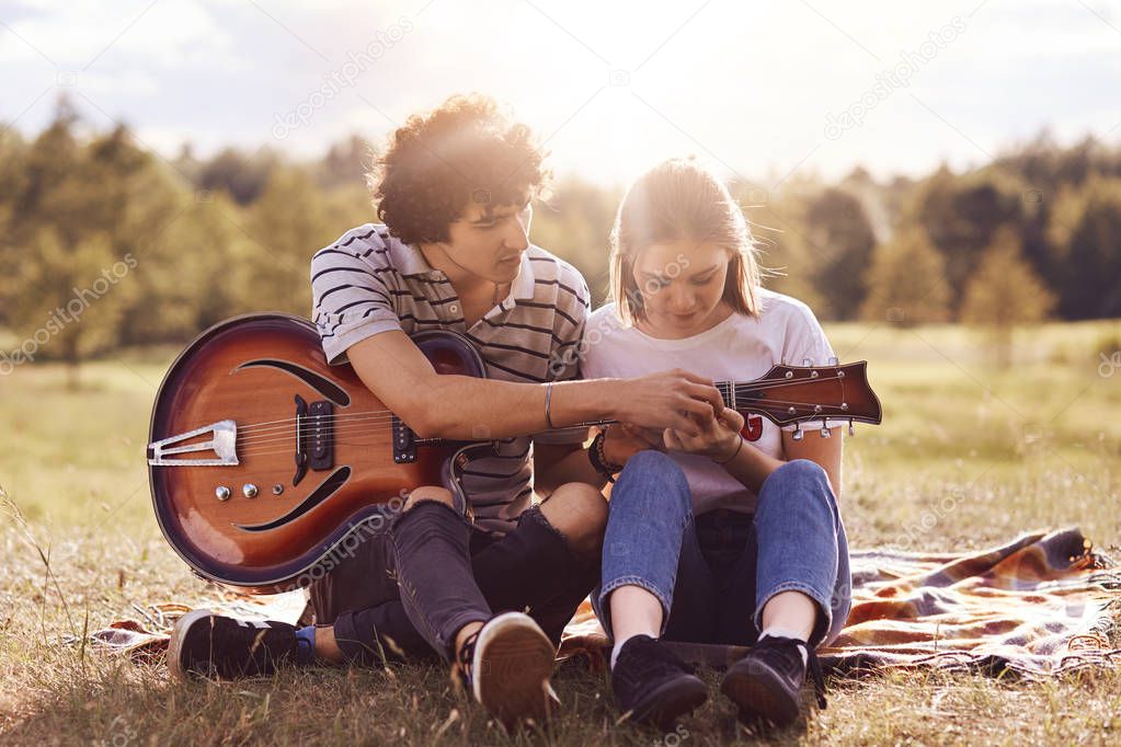 Picnic time. Lovely couple in love date on nature background, handsome male with curly hair teaches his beautiful girlfriend to play guitar, have good relationship. Dating, romance, lifestyle concept