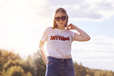 Şık tonları, t gömlek ve kot pantolon, pozlar gökyüzü Bankamatik karşı recreats standları açık yaz hava sırasında oldukça sevimli genç kadın. Şık genç tatil ve dinlenme zamanı vardır