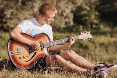 Çekici Avrupa erkek genç çimenlerin üzerinde oturur, çalış akustik gitar, recreats sırasında yaz günü, uygulamaları yeni şarkı, şarkı söylüyor, dışında sakin atmosferi tadını çıkarın. İnsanlar ve eğlence kavramı