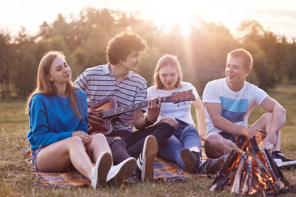 People and vacation concept. Cheerful best teenage friends enjoy romantic atmosphere on nature, have picnic together, play guitar, sit on plaid near campfire, have friendly talk with each other