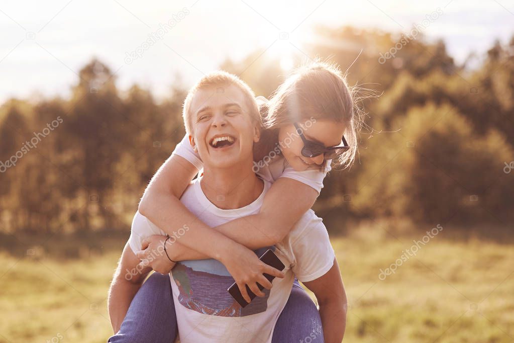 Funny happy boy gives piggyback to his girlfriend, foolish outdoor during summer day, demonstrate truthful feeling, have spare time during summer, enjoy togetherness. People and fun concept.