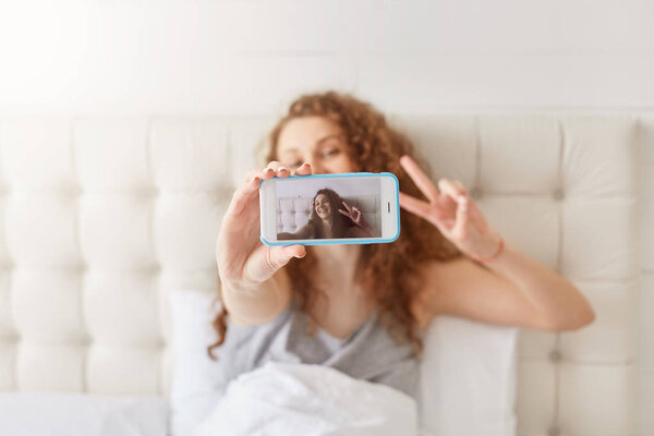 Female teenager poses for making selfie, makes peace sign, dressed in pyjamas, stays in bed during weekend, uses modern cellular, has satisfied expression. People, modern technology concept.