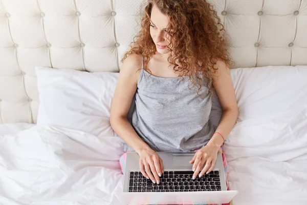 美丽的女人与卷曲的头发的顶端看着积极的一边 工作自由职业者在家里 键盘上的笔记本电脑 创造新的文章为杂志 摆在舒适的床上与白色亚麻布 — 图库照片