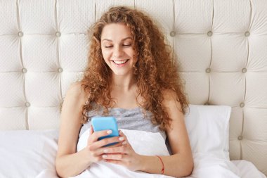 Neşeli genç güzel kadın bloggs sosyal ağlar, iletileri gönderir, beyaz keten ile rahat yatakta oturur, pozitif ifadesi olan, hafta sonu ve dinlenme odasında evde sahiptir