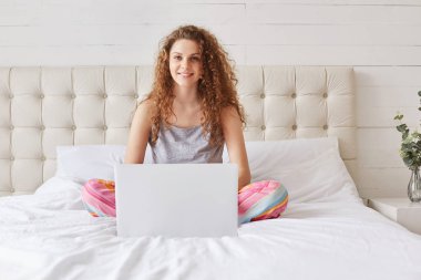 Positive beautiful female uses portable laptop computer for e learning, rests in domestic atmosphere at bedroom, dowloads files from networks or prepares for project. Woman in bedroom with device clipart