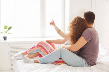 Arkadan görünüm sevgi çiftin tutkuyla kucaklamak, rahat yatağa otur ve pencereye bakın, gün ışığı, parmak ile bir şey erkek gösterdiğine tadını çıkarın. Evli kadın ve erkek yatak odasında