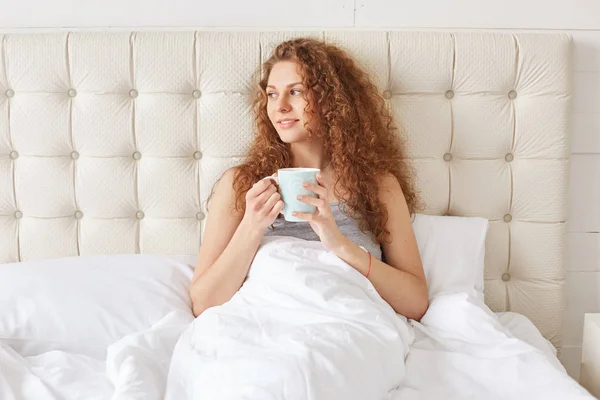 漂亮的年轻女人带着卷发在床上有晨香咖啡 看起来很惬意 享受日出 在卧室里有很好的放松 放松可爱的女性喝热茶独自在床上 — 图库照片
