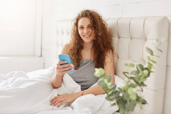 美丽的女人与卷曲的豪华头发享受在线沟通 在现代智能手机 坐在舒适的床上 醒来在良好的心情 正面女性使用电子设备在卧室里 — 图库照片