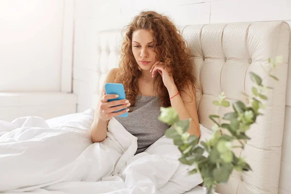可爱的年轻女子在睡衣 持有现代智能手机 阅读电子邮件表单朋友用心在移动应用 连接到无线互联网在卧室 花闲暇时间在卧室里 — 图库照片