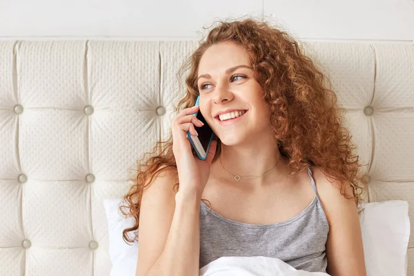 快乐无忧无虑的年轻女模特 卷曲的头发通过智能手机说话 Satisifed 听到好消息 花时间在床上 享受休息和沟通 床时间和技术概念 — 图库照片