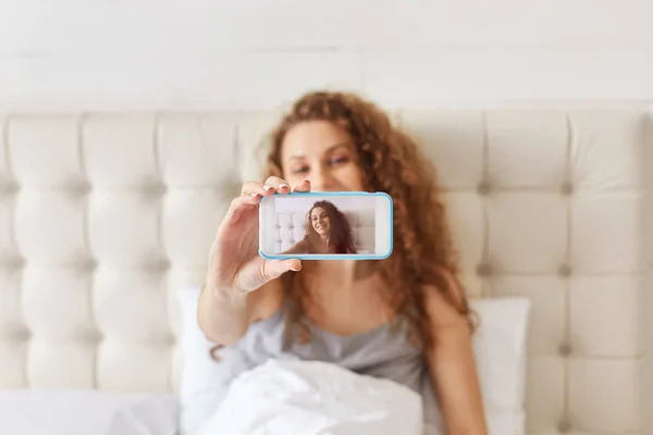 在床上 可爱的女人的肖像使自拍在智能手机上 看起来很高兴 并摆在相机 电话屏幕集中 漂亮的女性使用现代 Technlogies 拍摄自己 — 图库照片