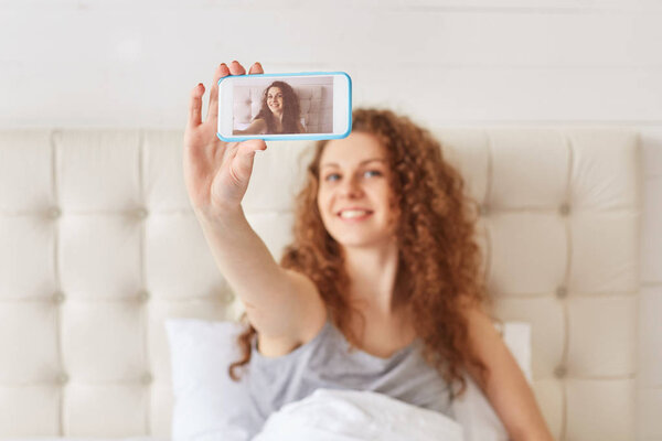 Photo of cheerful beautiful young woman with curly hair and pleasant smile, makes selfie on modern smart phone, going to share photos in social networks, rests in bedroom, has day off or weekend