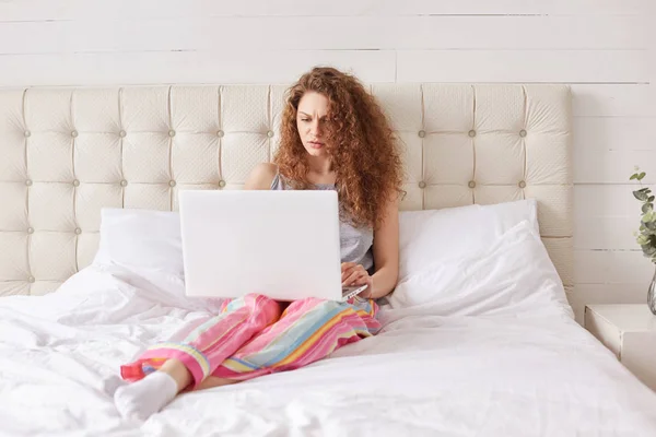 严肃的年轻女性自由职业者使用膝上型电脑为远程工作 在家中放松在床内部 穿戴随便 在网站上搜索信息 连接到无线互联网 — 图库照片