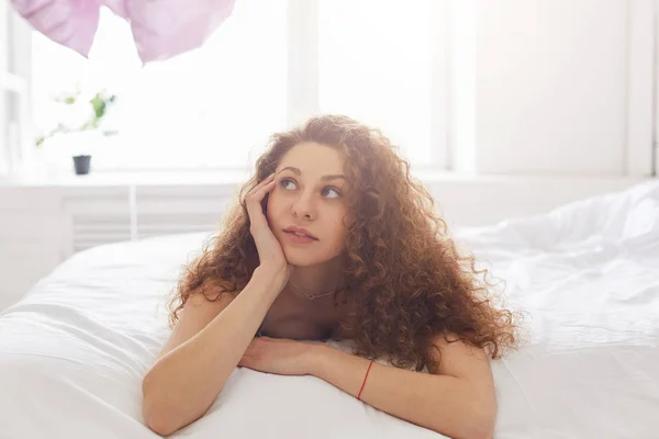 Indoor shot of dreamy woman with frizzy hair lies on bed with pensive expression, spends free time at bedroom, ponders about her plans for day. Beautiful female thinks about something before sleep