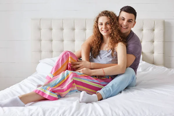 Young family couple sit and embrace on comfortable bed, enjoy togethernesss and good morning, have positive expressions, wear pyjamas. Curly beautiful woman spends free time in bedroom with lover