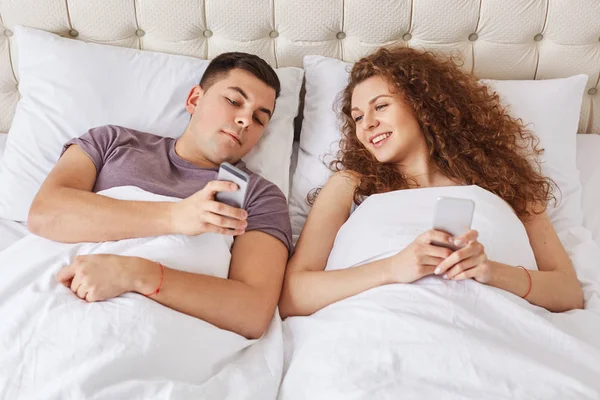 快乐深情的情侣在卧室里度过早晨的时光 总是保持联系 使用现代的小玩意上网和在线交流 家庭用移动电话在舒适的床 — 图库照片