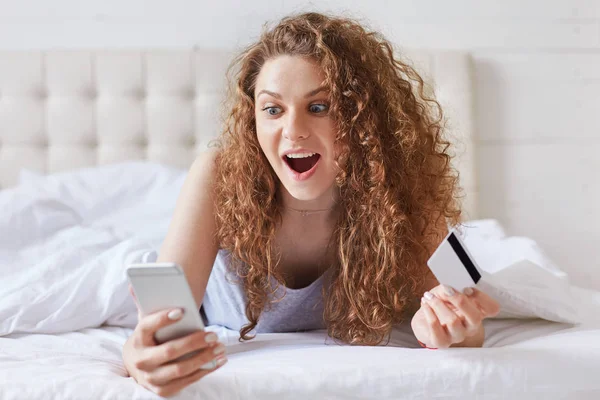 惊讶的兴奋女性卷曲的头发 检查网上帐户在智能手机 持有信用卡 使网上购物 而躺在舒适的床上 惊奇的年轻妇女喜悦新的购买 — 图库照片