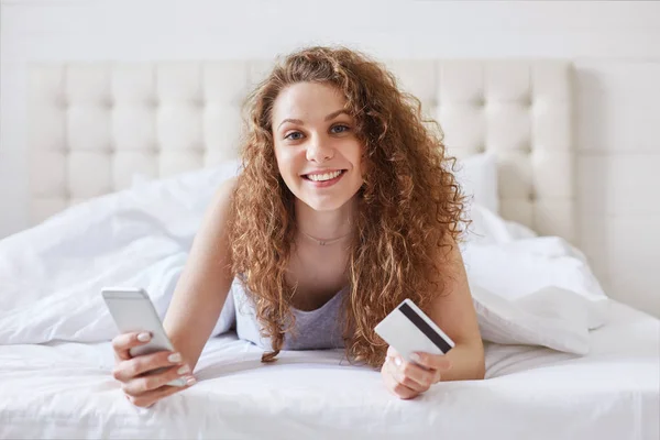 水平镜头可爱的女性穿着随意 感觉舒适的床上放松 持有现代手机和塑料卡 在线验证银行帐户 在家购物 支付购买 — 图库照片